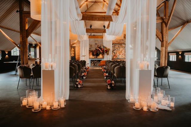 De Deel in ceremonieopstelling met groene stoelen, bloemen langs het looppad en witte doeken die over de houten plafondbalken zijn gedrapeerd.